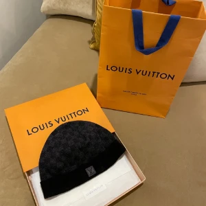 Svart mössa från Louis Vuitton - Säljer en snygg svart mössa från Louis Vuitton med klassiskt rutmönster. Mössan är i mycket bra skick och kommer i originalförpackning. Perfekt för höst och vinter, och den har en diskret LV-logga framtill. Passar både till vardags och lite finare tillfällen.