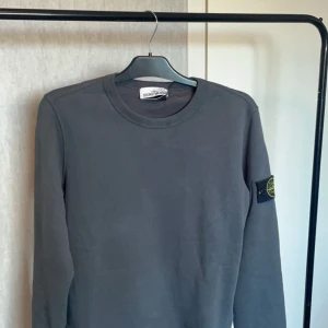 Grå tröja från Stone Island - Säljer en stilren grå tröja från Stone Island. Den har en klassisk rund hals och långa ärmar. På vänster ärm finns det ikoniska Stone Island-märket. Perfekt för både vardag och lite kyligare dagar. Passar bra till jeans eller chinos för en avslappnad look.