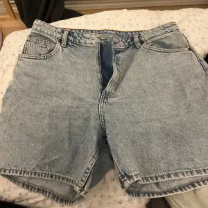 Säljer ett par klassiska blå jeansshorts från Monki i storlek 32. De har en normal passform med hög midja och är perfekta för sommaren. Shortsen har fem fickor och stängs med dragkedja och knapp. Perfekta för en avslappnad stil!