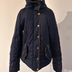 Mörkblå jacka med guldknappar - Säljer en snygg mörkblå jacka med guldknappar och dragkedjor. Den är quiltad och har praktiska fickor med dragkedjedetaljer. Perfekt för höst och vår!