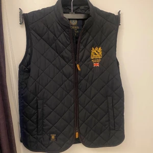 Svart quiltad väst från Morris - Säljer en snygg svart quiltad väst från Morris med dragkedja och broderad logga på bröstet. Perfekt för höst och vår. Västen har en stilren design med brittisk flagga och är i mycket bra skick.