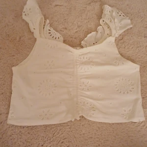 Vit ärmlös topp med spetsdetaljer - Säljer en superfin vit topp med spetsdetaljer och broderade blommönster. Den är ärmlös och har en söt volangkant vid axlarna. Perfekt för sommardagar eller en festlig kväll. Toppen är croppad och ger en luftig och bekväm känsla. Passar perfekt till jeans eller kjol!