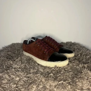 Vinröda sneakers från Lanvin - Säljer ett par snygga vinröda sneakers från Lanvin i mocka med svart tå och vit sula. De har snörning och är i mycket bra skick. Perfekta för både vardag och fest!