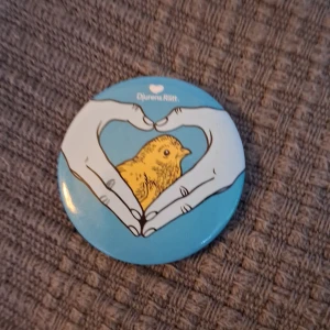 Djurens rätt pin med kycklingmotiv - En söt blå knapp med ett motiv av en gul kyckling inramad av händer som formar ett hjärta. Perfekt för att fästa på jackan eller väskan för att visa ditt stöd för djurens rättigheter. Knappen har en säkerhetsnål på baksidan för enkel fastsättning.