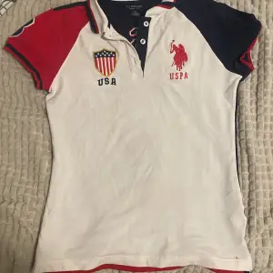 Säljer en snygg vit och marinblå pikétröja från U.S. Polo Assn. med röda detaljer. Tröjan har korta ärmar och broderade loggor fram och bak. Perfekt för en sportig look eller casual vardagsstil. Den är i bra skick har använt den en gång och redo för nya äventyr!