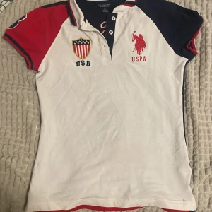Vit,marinblå och röd pikétröja från U.S. Polo Assn. - Säljer en snygg vit och marinblå pikétröja från U.S. Polo Assn. med röda detaljer. Tröjan har korta ärmar och broderade loggor fram och bak. Perfekt för en sportig look eller casual vardagsstil. Den är i bra skick har använt den en gång och redo för nya äventyr!