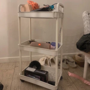 vit hylla - Vit rullvagn med tre hyllor fylld med olika föremål. På översta hyllan finns en rosa necessär och några håraccessoarer. Mellanhyllan har en liten låda och några oidentifierade föremål. På nedersta hyllan syns en svart låda och en hårborste. Perfekt för förvaring av smink och skönhetsprodukter.