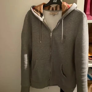 Grå hoodie från Burberry - Säljer en snygg grå hoodie från Burberry i mycket bra skick. Den har en dragkedja framtill och en klassisk Burberry-rutig insida i huvan. Perfekt för höst och vår, med en normal passform och långa ärmar. Superbekväm och stilren! Tveka inte på att fråga om du undrar något, den är självklart äkta, Storlek M men passar S också 