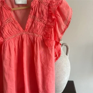 Korallfärgad blus från Neo noir - Säljer en superfin korallfärgad blus från Neo noir. Den har vackra volangdetaljer och spets vid ärmarna och halsen. Perfekt för vår och sommar! Blusen är i ett luftigt material som känns skönt mot huden.