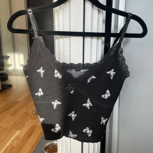 Svart linne med fjärilsmönster från HM - Säljer ett svart linne med vita fjärilar. Det har smala axelband och en v-ringad spetskant. Perfekt för sommardagar eller en avslappnad festkväll. Materialet är ribbat vilket ger en skön passform.