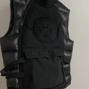 Svart dunväst från Glorious Gangsta - Säljer en svart dunväst från Glorious Gangsta i mycket bra skick. Västen har en quiltad design med dragkedja och ett stort märke på bröstet. Perfekt för höst och vinter, och ger en cool och stilren look. Passar bra till både vardag och fest.