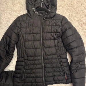 Svart Zara pufferjacka med huva - Säljer en svart pufferjacka i mycket bra skick. Den har en praktisk huva och dragkedja framtill. Perfekt för kalla dagar och håller dig varm och bekväm. Jackan har långa ärmar och två fickor med dragkedja.