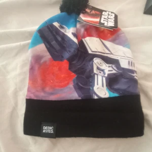 Star Wars mössa från Dedicated - Säljer en supercool Star Wars mössa från Dedicated! Den har ett färgglatt tryck med rymdmotiv i blått, rött och lila. Mössan är i nyskick och har en svart kant och en liten tofs på toppen. Perfekt för alla Star Wars-fans!