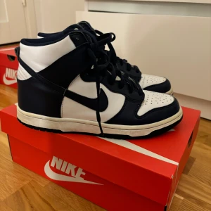 Nike Dunk High i svart och vit - Säljer ett par Nike Dunk High sneakers i klassisk marinblå och vit färgkombination. Skorna har en hög siluett med snörning och är i bra skick med några defekter.