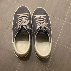 Philippe Model sneakers i blått och vitt - Säljer ett par Philippe Model sneakers i blått med vita detaljer. Skorna har en klassisk design med snörning och en bekväm passform. De är i välanvänt skick men fortfarande snygga och användbara. Perfekta för vardagsbruk!
