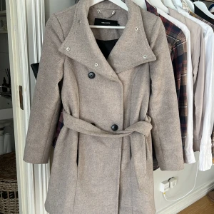 Beige kappa från Vero Moda - Säljer en snygg beige kappa från Vero Moda i storlek XS. Den har en klassisk design med knappar och ett bälte i midjan för en elegant look. Perfekt för höst och vår! Kappan är i välanvänt skick med lite noppor, men fortfarande superfin och användbar.