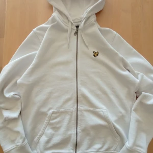 Vit Lyle & Scott hoodie med dragkedja - Säljer en vit hoodie med dragkedja från Yle & Scott. Tröjan har långa ärmar och en praktisk huva med dragsko. 