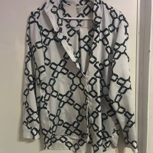 Vit och svart mönstrad blus - Snygg vit blus med svart kedjemönster. Blusen har långa ärmar och en klassisk krage. Perfekt för en stilren look.