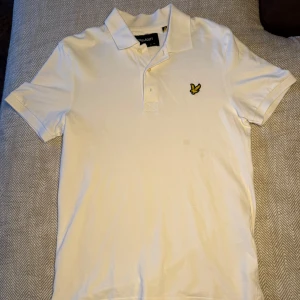 Vit pikétröja från Lyle & Scott - Säljer en stilren vit pikétröja från Lyle & Scott med korta ärmar och klassisk krage. Tröjan har en broderad logotyp på bröstet och knappar framtill. Perfekt för en avslappnad men snygg look.