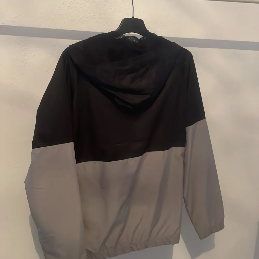 Säljer en stilren svart och grå vindjacka från Nike med dragkedja och huva. Jackan har långa ärmar och två praktiska fickor med dragkedja. Perfekt för blåsiga dagar. Nike-loggan är broderad på bröstet.. Takit.