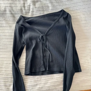 Svart ribbad topp med knyt - Snygg svart ribbad topp med långa ärmar och knyt framtill. Perfekt för en stilren look. Toppen har en croppad passform som ger en modern touch.