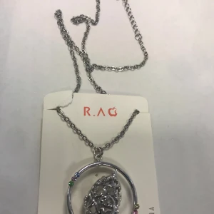 Silverfärgat halsband med dekorativt hänge - Elegant silverfärgat halsband från R.A.G med ett cirkulärt hänge. Hänget har en intrikat design med små färgade detaljer i grönt, blått, rött och gult. Perfekt för att ge en stilfull touch till din outfit. Helt ny 