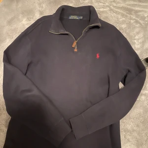 Svart långärmad tröja från Polo Ralph Lauren - Snygg svart långärmad tröja från Polo Ralph Lauren med dragkedja vid halsen och det klassiska röda logotypbroderiet på bröstet. Perfekt för en stilren och avslappnad look.