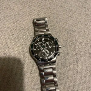 Swatch dark green irony YVS506G - Snygg silverfärgad klocka från Swatch med svart urtavla och vita detaljer. Klockan har en kronograf och en robust länkarmband i rostfritt stål. Perfekt för den som gillar en stilren och sportig look. Inga skador förutom små repor på armbandet, syns bara på nära håll. Kommer med extra lenkar och original låda🔥Priset kan självklart diskuteras🔥