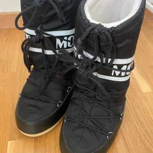 Svarta Moon Boots - Säljer ett par svarta Moon Boots med vit text och snörning. Perfekta för vintern med sin varma och vadderade design. De har en hög modell och är kända för sin unika stil och komfort.