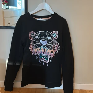 Svart sweatshirt från Kenzo - Säljer en svart sweatshirt från Kenzo med ett coolt tigertryck och texten 'Kenzo Paris' på framsidan. Tröjan har långa ärmar och en rund halsringning. Perfekt för en stilren och trendig look. Står att det är strl M men tycker att den sitter mer som en xs-s💓 Säljer pga att den inte kommer till användning, jätte snygg till ett par jeans.