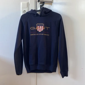 Mörkblå hoodie från GANT - Säljer en snygg mörkblå hoodie från GANT med tryck på framsidan. Tröjan har en klassisk känguruficka och en bekväm huva. Perfekt för en avslappnad stil. (Passar mig som bär xs/s)