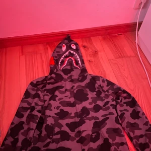 Bape tröja med grå svarta färger - Säljer en cool hoodie med camouflage-mönster och hajmotiv på huvan. Tröjan har dragkedja och är långärmad, perfekt för en streetwear-look.