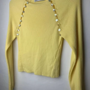 Gul ribbad tröja från Zara - Säljer en snygg gul ribbad tröja från Zara med långa ärmar och dekorativa knappar längs axlarna. Perfekt för att lysa upp garderoben med en pop av färg. Tröjan har en bekväm passform och är tillverkad i ett mjukt material.