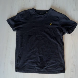 Svart t-shirt från Lyle & Scott - Säljer en stilren svart t-shirt från Lyle & Scott med deras ikoniska gula logga på bröstet. Perfekt för en casual look. T-shirten är kortärmad och gjord i mjuk bomull för extra komfort.