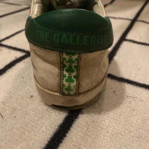 Vita sneakers med gröna detaljer från The Gallerist - Snygga vita sneakers med gröna detaljer och klövermotiv på sidan. Skorna har snörning och en grön hälflik med texten 'The Gallerist'. Perfekta för en avslappnad stil. Med kommer också en låda jag vill också påpeka att det inte finns kvar att köpa 