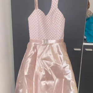 Rosa klänning från Erkut - Säljer en elegant rosa klänning från Erkut med ett glittrigt mönster på överdelen och en vacker satinliknande kjol. Klänningen har smala axelband och en dekorativ bältedetalj i midjan. Perfekt för speciella tillfällen! 🌸