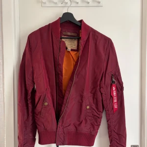 Vinröd bomberjacka från Alpha Industries - Snygg vinröd bomberjacka från Alpha Industries med dragkedja och ribbade muddar. Jackan har en klassisk design med fickor och en ikonisk röd tagg på ärmen. Perfekt för en stilren look. Nypris 1100kr