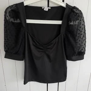 Svart topp från Amisu med prickiga ärmar - Snygg svart topp från Amisu med korta puffärmar i transparent tyg med prickigt mönster. Toppen har fyrkantig halsringning.