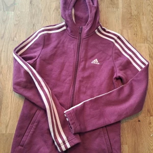 Vinröd huvtröja från Adidas - Säljer en vinröd huvtröja från Adidas med dragkedja och de klassiska tre ränderna längs ärmarna. Tröjan har en huva och är långärmad, perfekt för en sportig look. Adidas-loggan är broderad på bröstet.