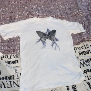 Vit t-shirt med fjärilstryck - Snygg vit t-shirt med ett stort fjärilstryck i svart och blått på framsidan. Perfekt för en avslappnad stil med en unik touch. Passar bra till jeans eller shorts.