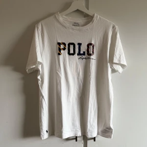 Vit t-shirt från Ralph Lauren - Säljer en klassisk vit t-shirt från Ralph Lauren med 'POLO' tryckt i färgglada bokstäver på bröstet. Den har en liten broderad logga i nederkant. Perfekt för en stilren look. XL i barnstorlek men sitter som s i vuxenstorlek. Knappt använd 