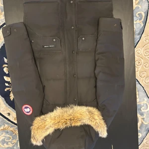 Svart parkajacka med päls från Canada Goose - Säljer en svart parkajacka från Canada Goose med pälsdetaljer på huvan. Jackan har långa ärmar och flera praktiska fickor framtill. Perfekt för kalla dagar med sin varma och skyddande design. 🖤