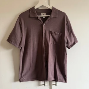 Lila kortärmad skjorta från H&M - Säljer en stilren lila kortärmad skjorta från H&M. Skjortan har en klassisk krage och knappar framtill samt en bröstficka. Perfekt för en avslappnad look. 