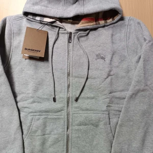  Burberry zip hoodie - Säljer en stilren grå hoodie från Burberry med dragkedja och broderad logga på bröstet. Tröjan har en justerbar huva med snören och praktiska fickor framtill. Perfekt för en avslappnad look.