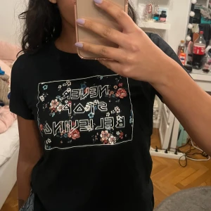 Svart t-shirt med blommigt tryck - Snygg svart t-shirt med texten 'Never Stop Believing' i ett blommigt mönster på framsidan. Perfekt för en casual look med en touch av stil. Kortärmad och bekväm att bära.