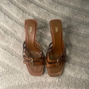 Bruna sandaler från Zara - Snygga bruna sandaler från Zara med två remmar och spännen. Perfekta för sommarens varma dagar. Sandalerna har en fyrkantig tå och en låg(7,5 cm) klack för extra komfort.        Använt få timmar en gång.                                         •	Yttermaterial: 100 % kohud.                                       •	Foder: 100 % getskinn.                                             •	Sula: 100 % termoplastiskt polyuretan.                 •	Innersula: 85 % getskinn, 15 % termoplastiskt polyuretan