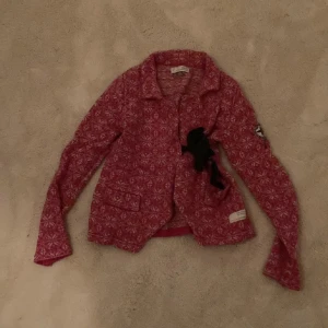 Rosa mönstrad cardigan med broderi - Säljer en unik rosa cardigan med ett snyggt mönster och broderad kompassdetalj på ärmen. Tröjan har långa ärmar och en dekorativ rosett framtill. Perfekt för att ge din outfit en färgglad touch!