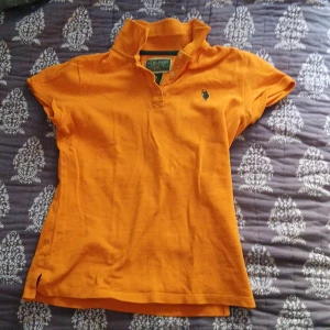 Orange pikétröja från US Polo Assn - Säljer en snygg orange pikétröja från US Polo Assn med korta ärmar och klassisk krage. Tröjan har en broderad logga i blått på bröstet och knappar vid halsen. Perfekt för en avslappnad stil!