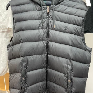 Svart Ralph lauren väst  - Snygg svart dunväst från Ralph Lauren med dragkedja och broderad logga på bröstet. Västen har en praktisk huva och två fickor framtill med knappar. Perfekt för kyligare dagar. BVSA. Storlek L men sitter mer som M