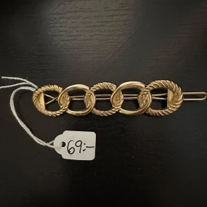 Guldfärgad hårklämma med ringar - Elegant guldfärgad hårklämma med fem sammanlänkade ringar i ett repmönster. Perfekt för att ge en stilfull touch till din frisyr. Klämman är cirka 8 cm lång och har en säker låsmekanism.